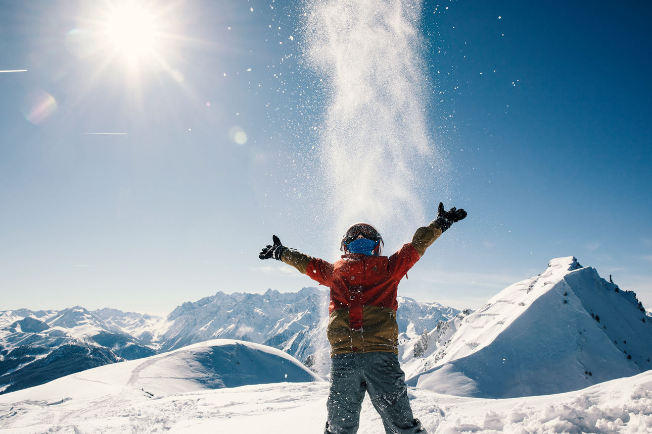 A young boy throwing snowballs in the bright sunshine, with a blue ski and snow capped mountain peaks in the background. Taken in Verbier, Switzerland.