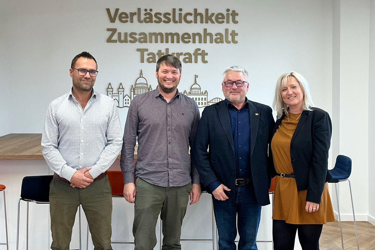 Drei Männer und eine Frau stehen für ein Foto v or einer Wand, auf der Verlässlichkeit, Zusammenhalt und Tatkraft steht