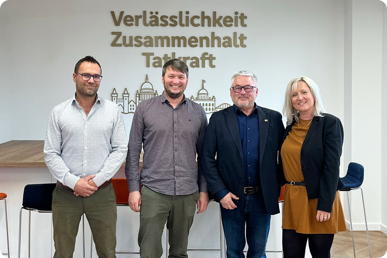 Drei Männer und eine Frau stehen für ein Foto v or einer Wand, auf der Verlässlichkeit, Zusammenhalt und Tatkraft steht