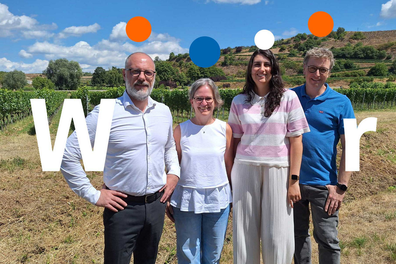 „Ein vierköpfiges Team steht für die ‚Wir‘-Kampagne in einem Weinberg. Die ländliche Umgebung unterstreicht die Werte Gemeins