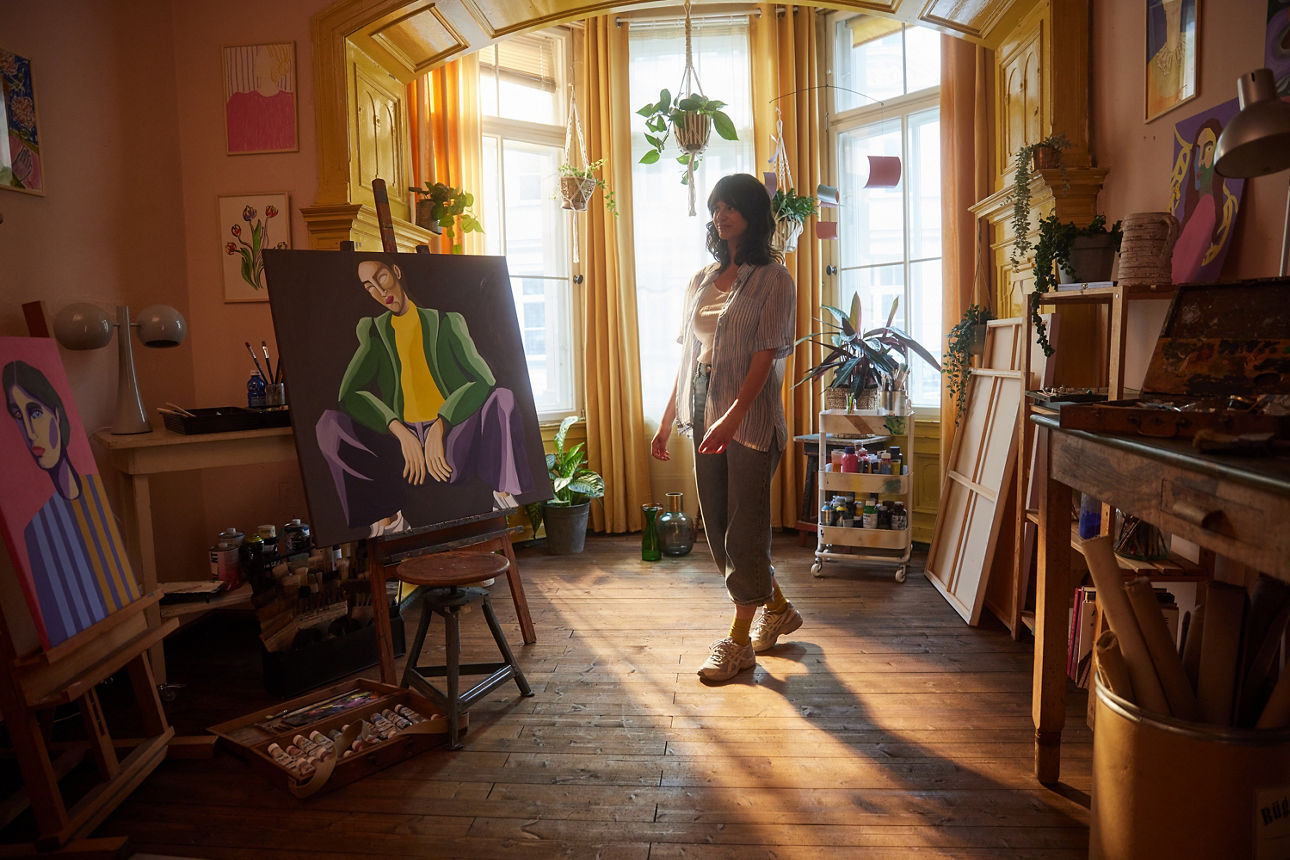 Eine junge Frau steht mitten in ihrem Atelier. Im Hintergrund ist eine helle Fensterfront. Zu sehen sind außerdem ein großes Gemälde, sowie viele Malutensilien im Raum. Eine junge Frau steht mitten in ihrem Atelier. Im Hintergrund ist eine helle Fensterfront. Zu sehen sind außerdem ein großes Gemälde, sowie viele Malutensilien im Raum.