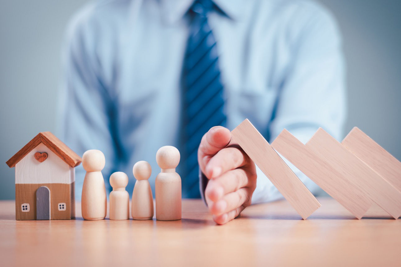 Ein Mann mit Krawatte schützt Holzfiguren und ein Holzhaus mit seiner Hand vor umfallenden Holzbausteinen