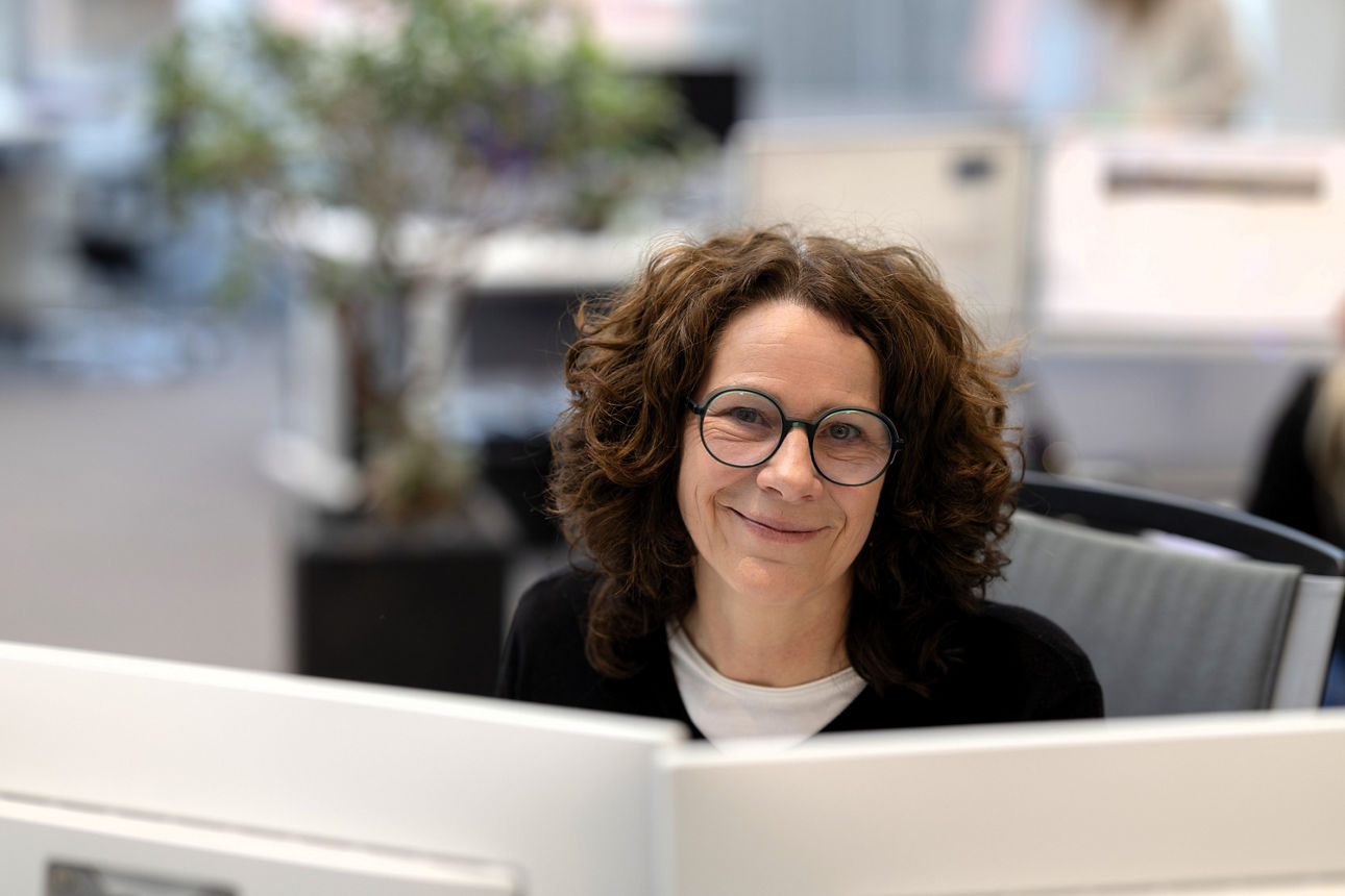 Eine weibliche Person sitzt in einem modernen Büro an einem Arbeitsplatz hinter mehreren Monitoren. Im Hintergrund stehen Bür