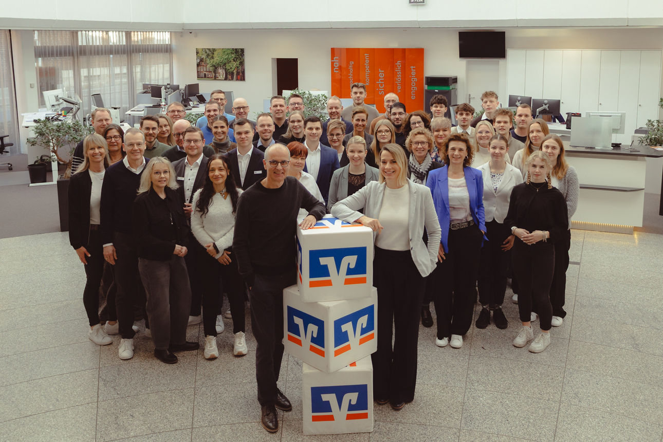 Eine größere Gruppe von Personen steht in einer offenen, hellen Eingangshalle zusammen. Im Vordergrund stehen drei Würfel mit