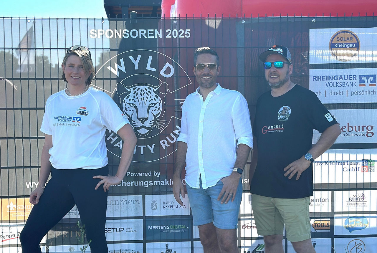 Drei Personen stehen vor einem Zaun mit dem Logo 'WYLD Charity Run 2025'. Die Person links trägt ein weißes T-Shirt, die mittlere Person ein weißes Hemd und die rechte Person ein schwarzes T-Shirt.