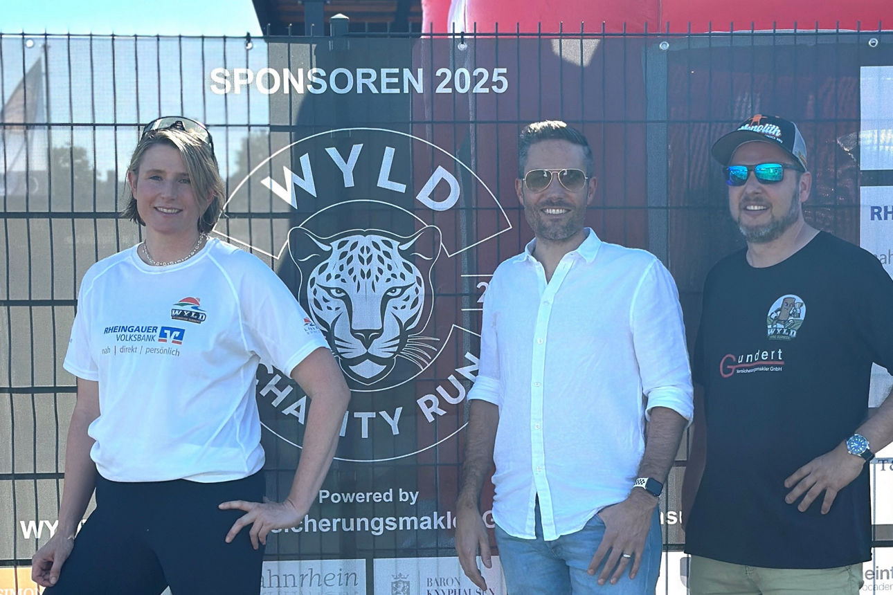 Drei Personen stehen vor einem Zaun mit dem Logo 'WYLD Charity Run 2025'. Die Person links trägt ein weißes T-Shirt, die mittlere Person ein weißes Hemd und die rechte Person ein schwarzes T-Shirt.