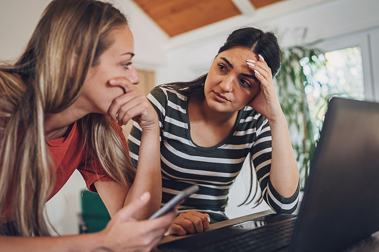Zwei Frauen stehen mit den Ellenbogen abgestützt vor einem Tisch. Darauf steht ein geöffneter Laptop. Eine der beiden hält ein Handy in der Hand. Beide wirken verzweifelt und setzen sich mit einem Problem auseinander.