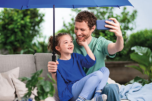 Vater und Tochter unter einem blauen Schirm auf einem Sofa im Freien machen ein Selfie
