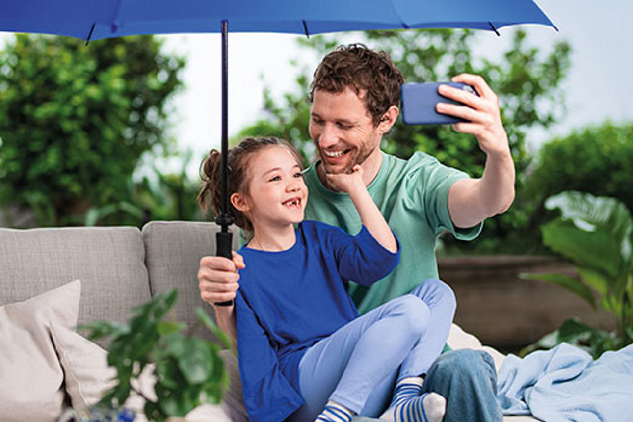 Vater und Tochter unter einem blauen Schirm auf einem Sofa im Freien machen ein Selfie