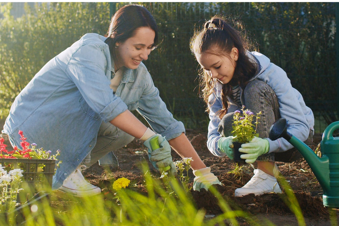 Zwei Personen arbeiten gemeinsam im Garten und pflanzen Blumen, daneben eine grüne Gießkanne und ein orangefarbener Hinweis.