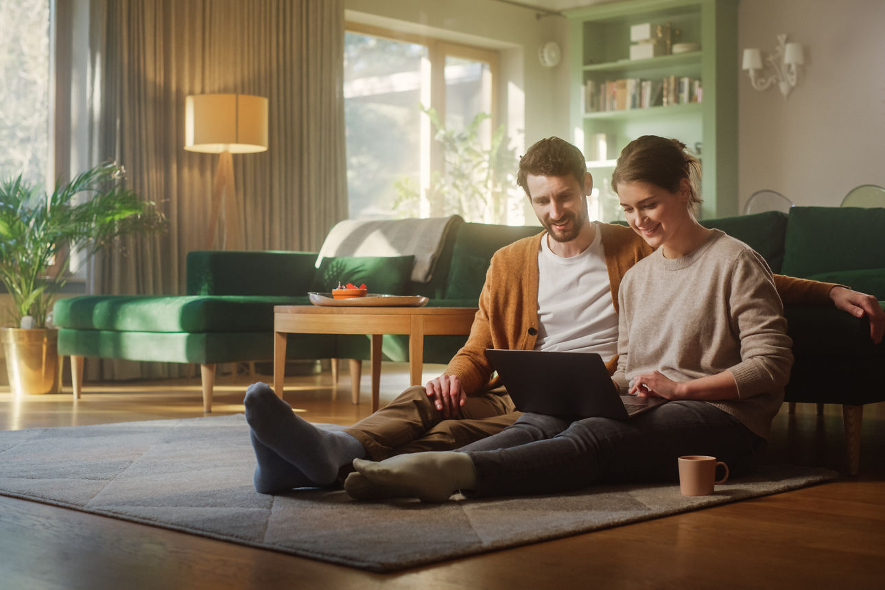 Ein Paar sitzt auf dem Boden vor der Couch und schaut gemeinsam auf Laptop