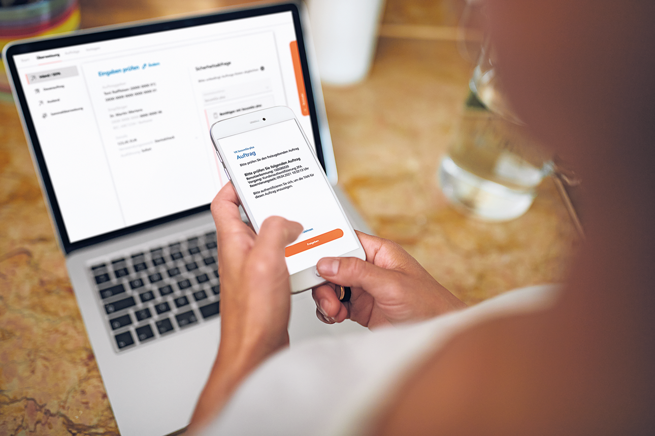 Eine Frau sitzt vor einem aufgeklappt Laptop auf dem das OnlineBanking zu sehen ist und bestätigt eine Transaktion mit dem Smartphone