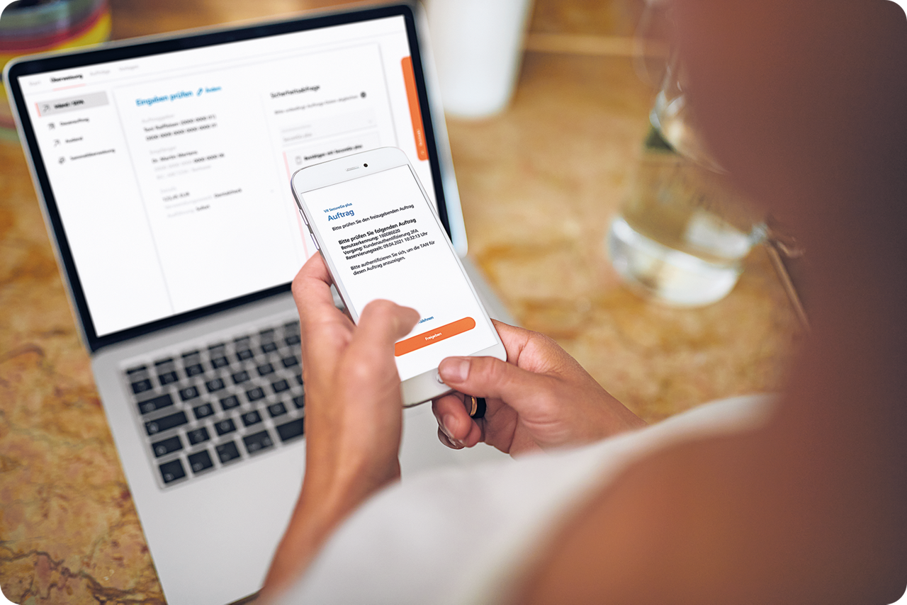 Eine Frau sitzt vor einem aufgeklappt Laptop auf dem das OnlineBanking zu sehen ist und bestätigt eine Transaktion mit dem Smartphone