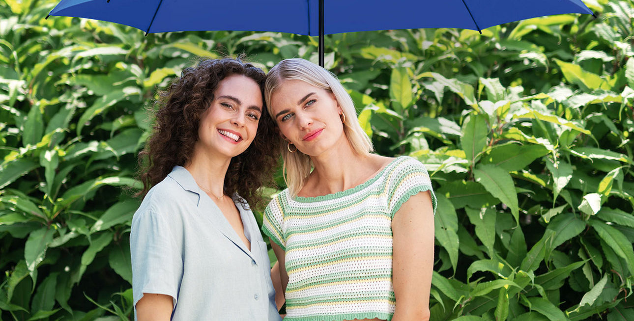 Zwei junge Frauen unter einem blauen Schirm stehen vor einer Hecke