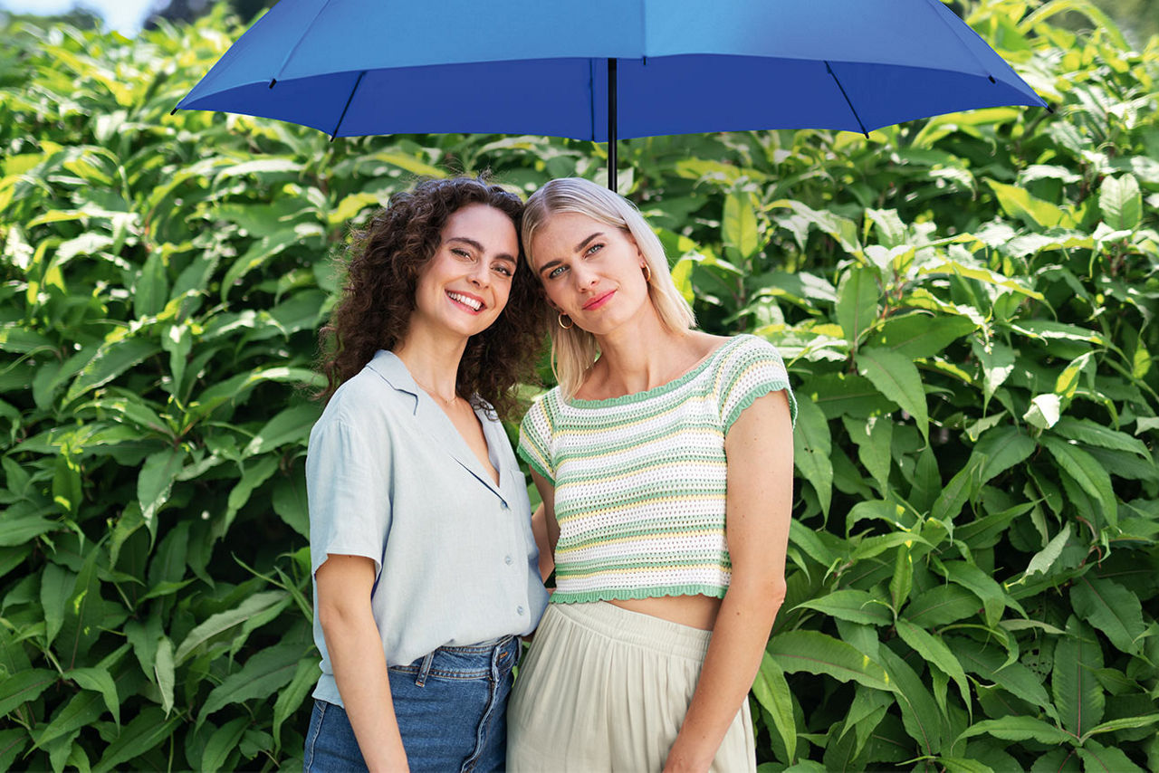 Zwei junge Frauen unter einem blauen Schirm stehen vor einer Hecke