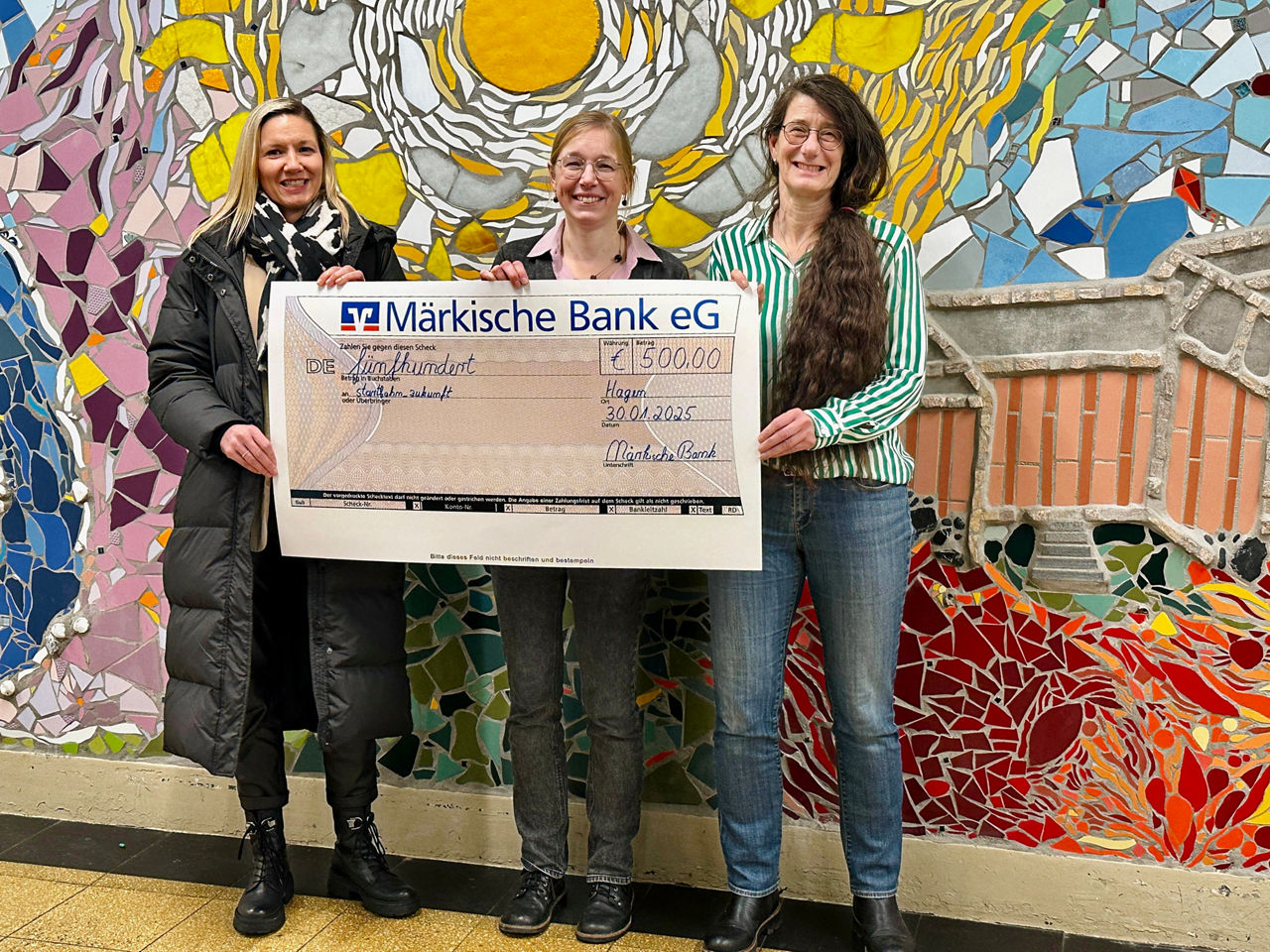 3 Personen von Märkischer Bank, Agentur Mark und Sekundarschule Altenhagen vor bunter Wand mit Zuwendungsscheck in der Hand.